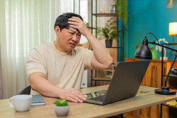 Asian man sitting at home table facepalms after reading absurd news on laptop, eyes shut in...