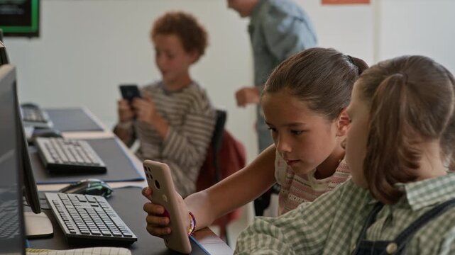 Two cute schoolgirls sitting in classroom and looking at smartphone screen while watching online video against happy boys discussing curious stuff in mobile gadget