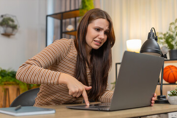 Young woman at home sees laptop freeze with error and virus alert waves hand frustrated and...