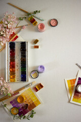 Flat lay of colorful watercolor palette, brushes, jars of paint, floral elements, and painted swatches on paper. Artistic workspace background, perfect for creativity, design projects, and inspiration