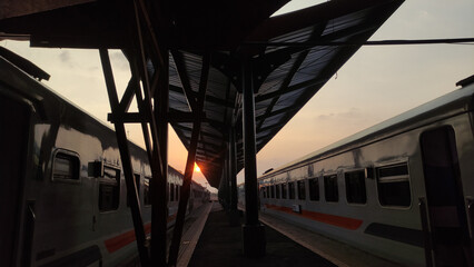 A railway station view at sunset with two trains on both sides of the platform. The warm sunlight...