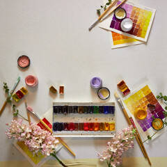 Flat lay of colorful watercolor palette, brushes, jars of paint, floral elements, and painted swatches on paper. Artistic workspace background, perfect for creativity, design projects, and inspiration
