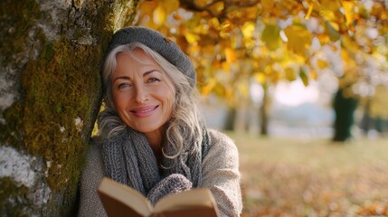 a medium shot of a happy senior woman relaxing under a tree while holding a book in an autumn park no logos no brands ar 169