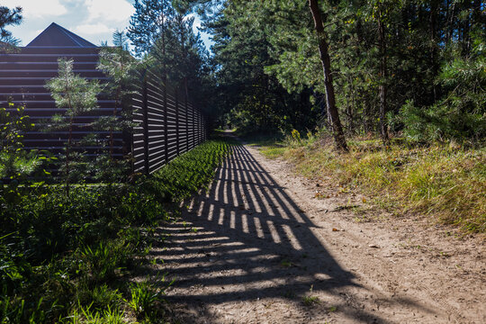 a path running along a high horizontal dark-colored fence. Bright rays of sun break through the fence, casting long, parallel shadows on the path and grass.