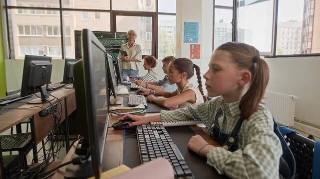 Serious schoolgirl looking at computer screen and clicking mouse, then turning her head, listening to teacher and raising hand with the rest of class at lesson of programming