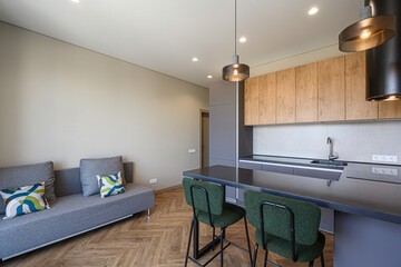 Modern kitchen and living area with gray sofa, wood cabinets, bar stools, and stylish lighting.  Bright, minimalist design