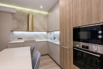 modern kitchen with neutral tones. Features wooden cabinets, marble backsplash, a sleek oven, and a white table