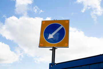 Road sign with blue circle and white arrow pointing downwards, indicating direction on street. Reflective orange dots enhance visibility for drivers, ensuring safe traffic flow