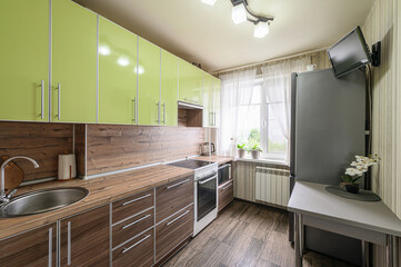 Cozy kitchen with green cabinets, wood countertops, and a stainless steel sink. A TV is mounted on the wall near a refrigerator
