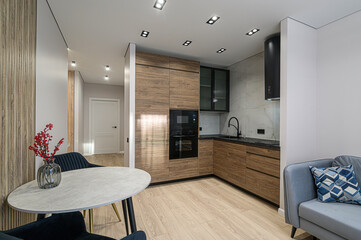 Modern kitchen with wood cabinets, black appliances, a round dining table, and a gray sofa. Well-lit and stylishly designed