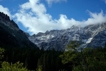 Obraz premium Snow-dusted rocky peaks towering above evergreen forest with drifting clouds under clear blue sky, forming dramatic mountain scenery.