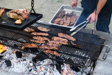 Korean style beef cooking on an outdoor barbeque grill with hot coal below, chef using tongs to remove cooked beef and place in container, as part of luau dinner preparations
