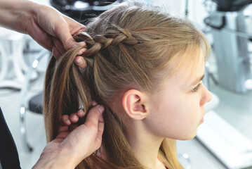 Fototapeta premium Hairdresser weaves a braid to a preteen blond girl in a beauty and hair salon