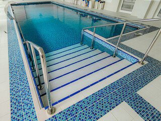 Clean indoor swimming pool with blue mosaic tiles, stainless steel handrails, and shallow entry steps, designed for training, therapy, or leisure.