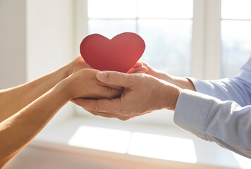 Two pairs of hands gently holding red paper heart together. Symbol of love, care, romance, charity,...