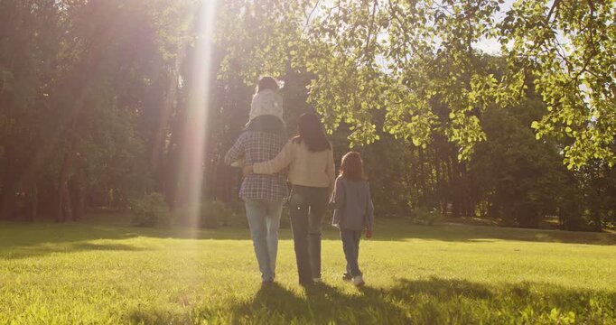 Happy family walk in summer park, mother and father, sibling kids walking together in sunny day of weekend. Rear view of mom hugging dad and holding sons hand, man carrying daughter on shoulders