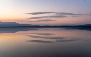 A serene lake reflects the pastel hues of a twilight sky, with silhouetted mountains in the distance and wispy clouds creating a tranquil and breathtaking natural landscape scene