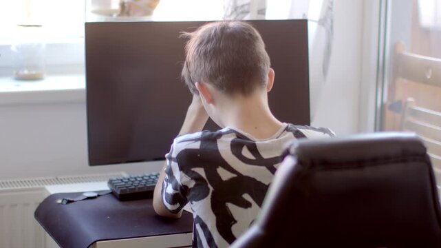 A young child shows visible signs of frustration and disappointment while sitting at a desk with a computer in front of him
