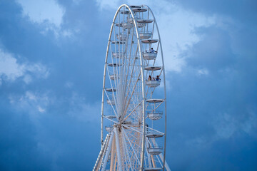 A towering Ferris wheel with enclosed gondolas rises against a dramatic blue sky filled with scattered clouds in Bulgaria