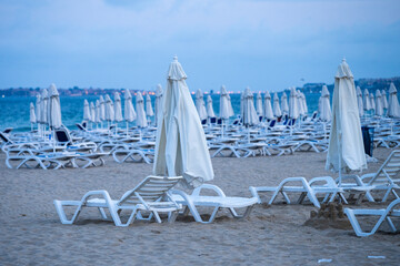 Rows of empty white lounge chairs and umbrellas line a quiet, overcast beach with a distant...