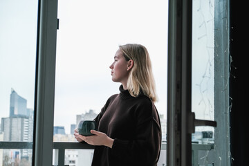 Mindful city morning: a woman on a balcony in a cozy sweater holds a warm mug, pausing between home...