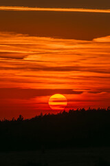 A fiery sunset glows behind a silhouetted treeline, painting the sky with vibrant shades of orange, red, and gold.