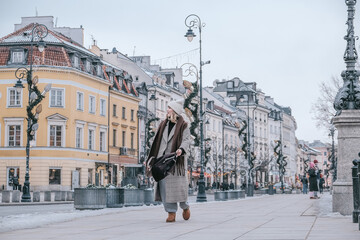 Winter city wanderlust: female tourist walking historic Warsaw old town, pastel facades and holiday lights, cozy coat, scarf and beanie. Urban sightseeing, slow travel, sustainable Europe escape eco.