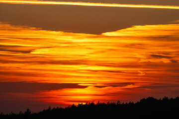 A brilliant sunset blazes behind a forested horizon, illuminating layered clouds in fiery tones of orange, red, and gold.