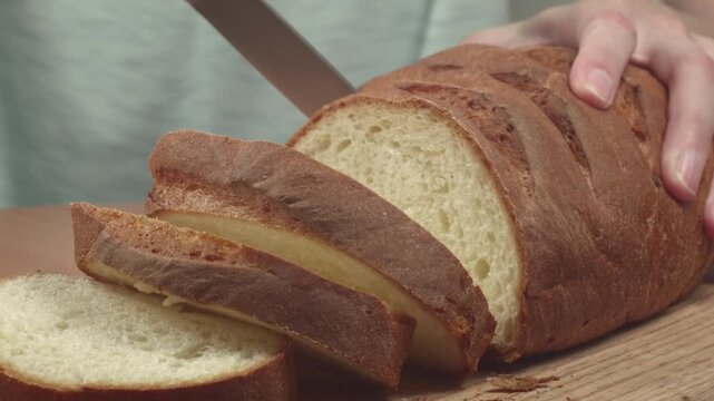 Close-up of young woman slicing fresh white bread with a kitchen knife on a cutting board in her kitchen, recorded with ambient sound.