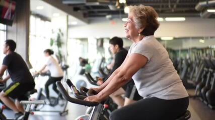 A senior group of individuals, including a joyful elderly Latina woman, engages in a lively cycling session at a modern gym. Bright lights and a welcoming atmosphere enhance their workout experience - Powered by Adobe