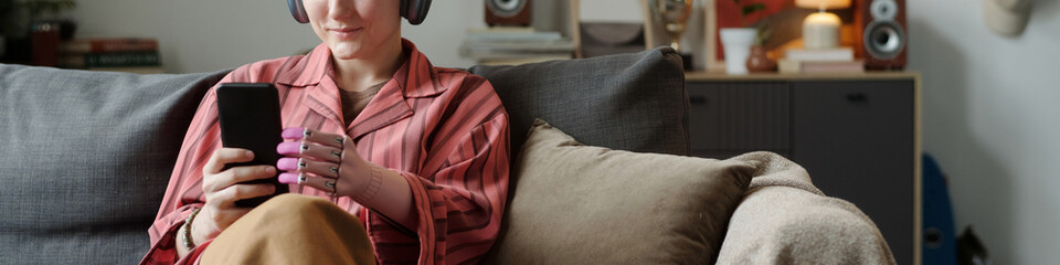 Caucasian young adult woman with prosthetic hand sitting on sofa wearing headphones using...