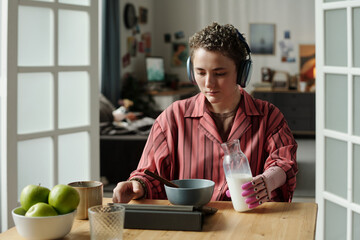 Young adult Caucasian woman with prosthetic hand wearing headphones sitting at table using digital tablet while pouring milk into bowl, demonstrating gadget addiction during breakfast