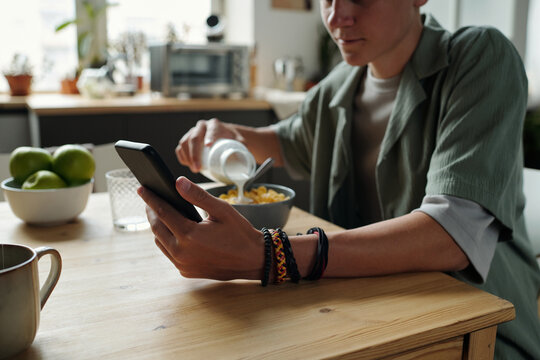 Young Caucasian man sitting at kitchen table holding smartphone in one hand while pouring milk into bowl of cereal with other hand, multitasking during breakfast, showing gadget addiction