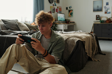 Caucasian teenage boy sitting on floor intensely playing handheld gaming console in bedroom,...