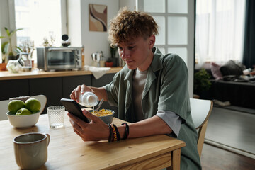 Caucasian teenage boy sitting at kitchen table using smartphone while eating breakfast cereal, focusing on screen and holding spoon, demonstrating gadget addiction during meal
