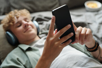 Caucasian teenage boy lying on bed using smartphone wearing headphones smiling while engaging with...