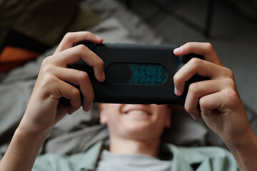 Teenage Caucasian boy lying on bed holding smartphone with both hands playing mobile game smiling showing signs of gadget addiction