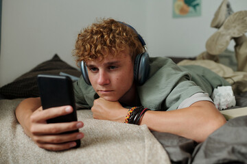 Caucasian teenage boy lying on bed using smartphone wearing headphones showing signs of gadget...