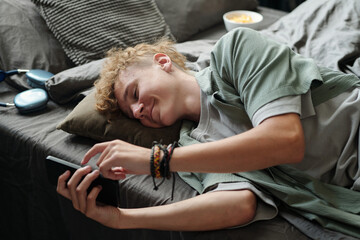 Caucasian teenage boy lying on bed using smartphone with headphones nearby showing signs of gadget addiction, focusing intently on screen while smiling and interacting with device