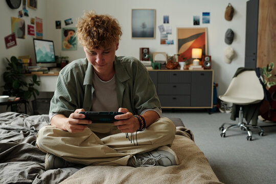 Teenage Caucasian boy sitting cross legged on bed intensely playing handheld gaming console in modern bedroom surrounded by personal items illustrating gadget addiction concept