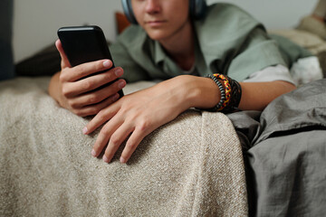 Teenager boy with light skin lying on bed using smartphone wearing headphones focusing on screen...