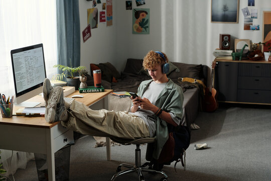 Teenage Caucasian boy sitting in messy bedroom wearing headphones, leaning back with feet on desk, intensely using smartphone while ignoring computer and study materials nearby