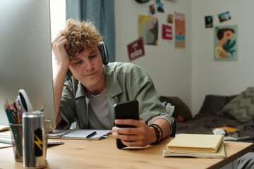 Caucasian teenage boy wearing headphones sitting at desk holding smartphone with distracted...