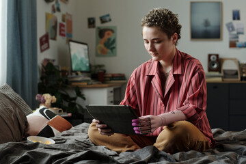 Young adult Caucasian woman with prosthetic hand sitting on bed using digital tablet, focusing...