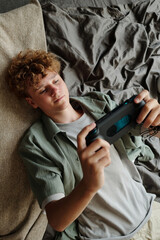 Teenage Caucasian boy lying on sofa intensely playing handheld gaming console, focusing on screen,...