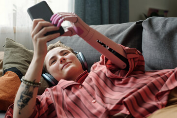 Caucasian young adult woman with prosthetic arm lying on sofa using smartphone wearing headphones...