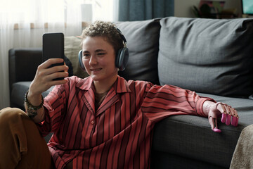 Caucasian teenager sitting on floor using smartphone with headphones, smiling while engaging with...