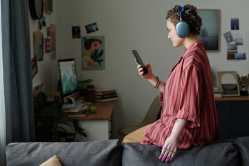 Young adult Caucasian woman with disability wearing headphones sitting on sofa using smartphone...