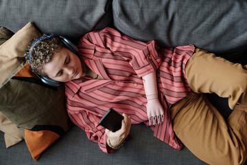 Woman with prosthetic arm lying on couch wearing headphones holding smartphone staring at screen...