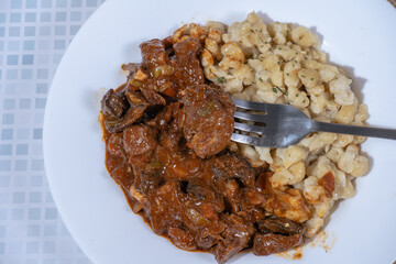 Fork taking a bite of beef goulash with spaetzle on white plate